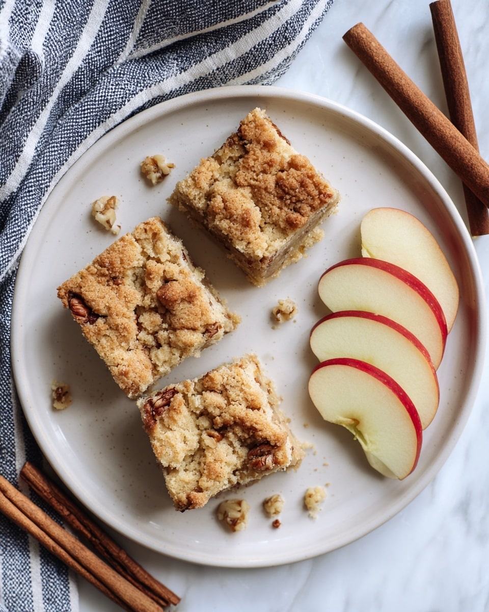 A white plate shows three golden brown squares of crumbly cake with visible nut pieces on top, arranged side by side on the left. To the right, there are five thin slices of red-edged apple fanned out neatly. Small nut crumbs are scattered around the plate. The plate is set on a white marbled surface, next to two brown cinnamon sticks and a blue and white striped cloth. Photo taken with an iphone --ar 4:5 --v 7