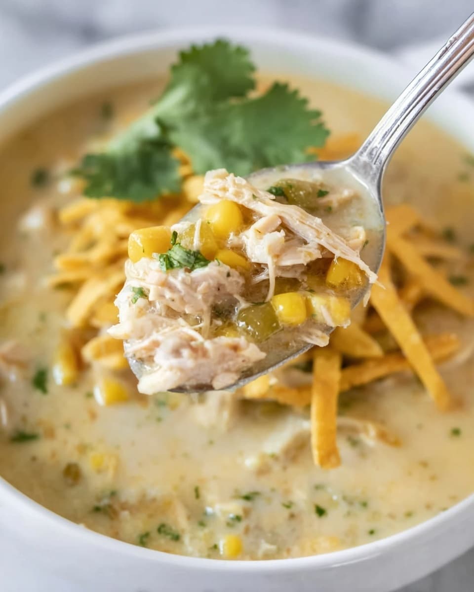 A close-up of a spoon holding creamy soup with visible layers of shredded chicken pieces, yellow corn kernels, small green herbs, and a thick, smooth, light beige broth. In the background, the soup is in a white bowl filled with the same creamy mixture and topped with crispy golden yellow strips. There is a green cilantro leaf resting on the soup near the back of the bowl. The bowl sits on a white marbled surface. The colors blend softly with the textures showing smooth and chunky elements photo taken with an iphone --ar 4:5 --v 7