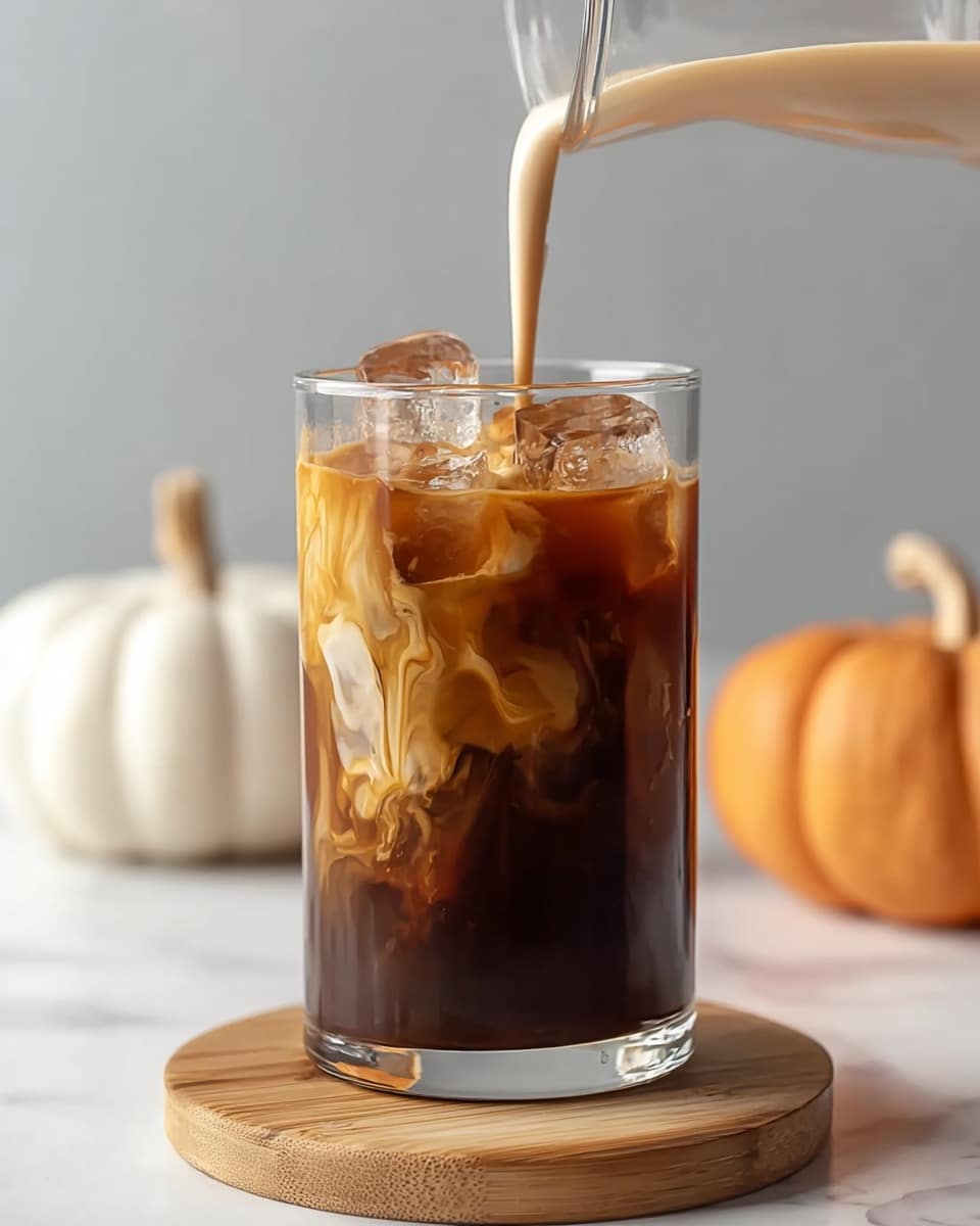 A clear glass filled with dark brown iced coffee and large ice cubes, placed on a round wooden board set on a white marbled surface. Light cream is being poured from a glass container into the coffee, creating a swirling, foamy light brown layer on the top edge with some cream turning lighter shades as it mixes with the dark coffee below. In the background, out of focus, there are two pumpkins, one white and one orange, against a plain light gray background. Photo taken with an iphone --ar 4:5 --v 7