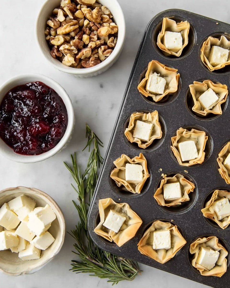 A dark gray muffin tray filled with small pastry cups, each holding a square piece of creamy white cheese. The pastry cups are light golden beige with uneven, crinkled edges. To the left of the tray are three white bowls on a white marbled surface: the top bowl contains mixed nuts of various brown shades, the middle bowl is filled with dark red jam with pieces in it, and the bottom bowl has extra pieces of the same white cheese. At the bottom left corner, there is a sprig of fresh green rosemary. The overall scene is bright and clean. photo taken with an iphone --ar 4:5 --v 7