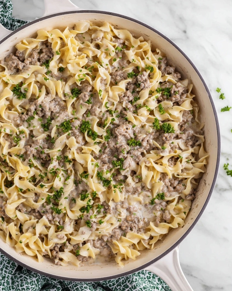 The image shows a white pan filled with creamy egg noodles mixed with ground beef. The dish has roughly two layers: a base of soft, pale yellow egg noodles with a smooth texture, and evenly spread cooked ground beef mixed throughout in a light brown color. The creamy sauce lightly coats the noodles and meat, giving the whole dish a moist, glossy look. Small green parsley pieces are scattered on top, adding a touch of color. The pan is sitting on a white marbled surface with a green and white patterned cloth nearby. Photo taken with an iphone --ar 4:5 --v 7