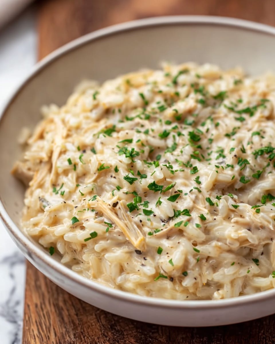 A white bowl filled with a creamy dish made of soft, small rice grains mixed with shredded light brown chicken pieces. The creamy sauce has a smooth texture and light beige color that lightly coats the rice and chicken. The top is sprinkled with small bits of green herbs, adding a fresh pop of color over the warm and hearty main layer. The bowl is placed on a white marbled surface with a subtle pattern. photo taken with an iphone --ar 4:5 --v 7