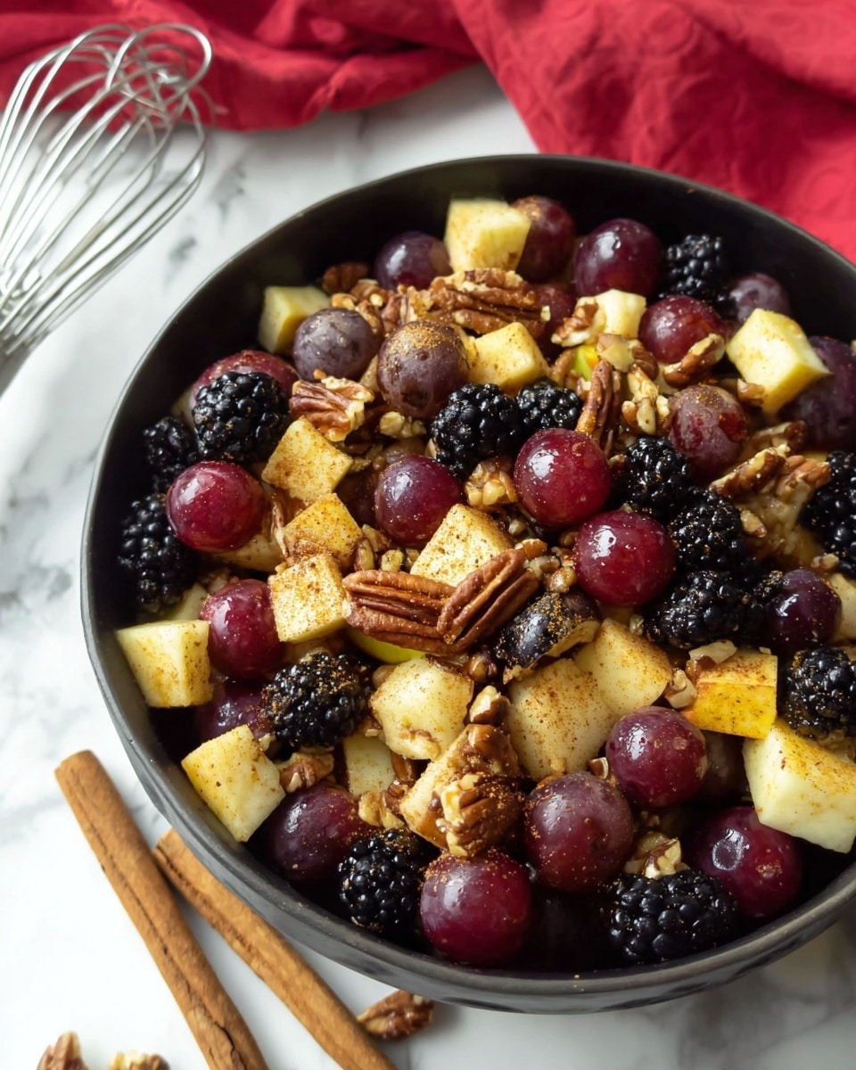 The image shows a close-up of a dark bowl filled with a colorful fruit and nut mix. The top layer consists of shiny red grapes, black blackberries, and small chunks of yellow apple dusted with a light brown spice, likely cinnamon. Scattered among the fruit are whole and chopped pecans adding a rough texture. The bowl is placed on a white marbled surface with two cinnamon sticks nearby and a metal whisk and red cloth in the background. The colors are rich and natural, highlighting the freshness of the fruit and the crunchiness of the nuts. photo taken with an iphone --ar 4:5 --v 7
