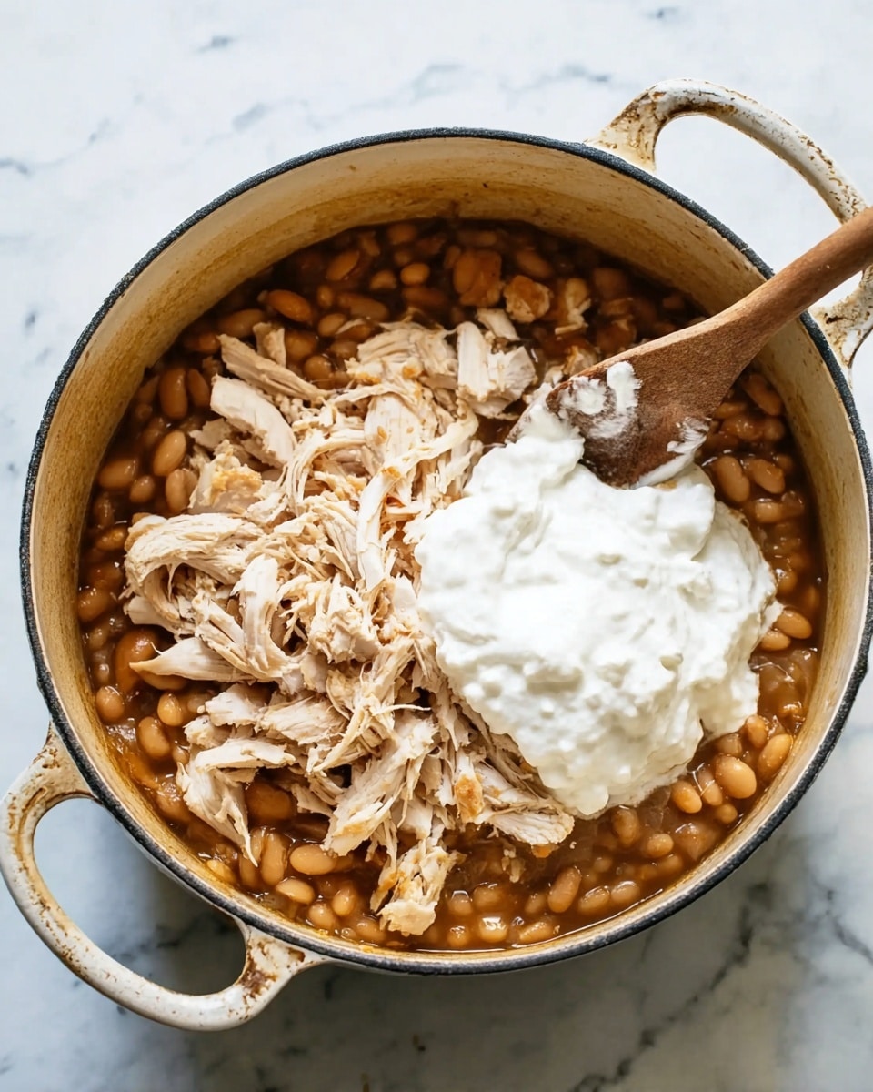 The image shows a white pot on a white marbled surface, filled with three main layers: a base layer of cooked beans in a brown sauce with a soft texture, on top of that shredded light-colored chicken, and a dollop of thick, white cream layered in the center on top of the chicken. A wooden spoon rests inside the pot, angled toward the left side. The pot has two handles and a rustic appearance. The photo is taken with an iphone --ar 4:5 --v 7