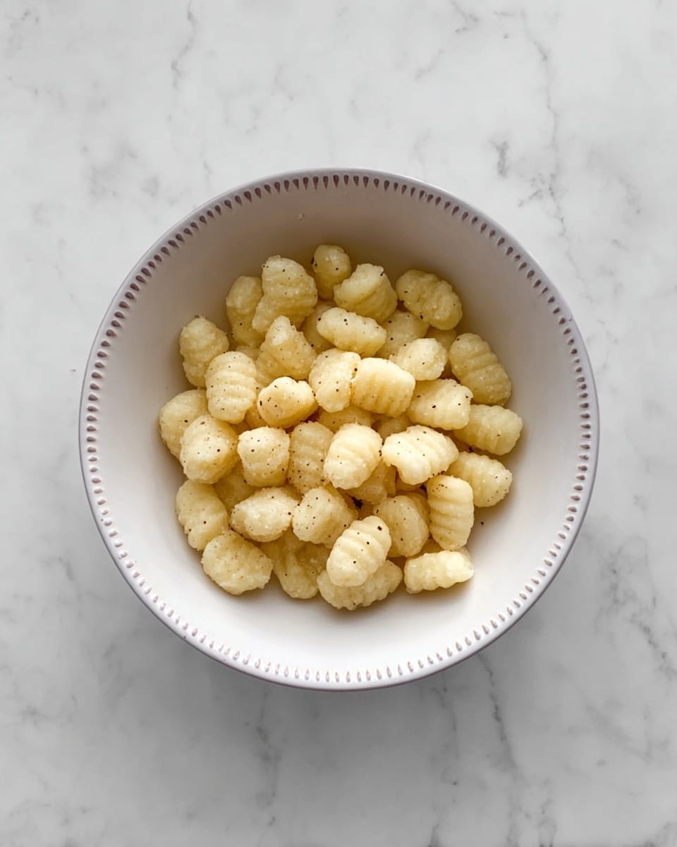 The image shows a white bowl filled with a single layer of small, soft gnocchi pieces. Each gnocchi is pale yellow with a slightly rough texture and some visible black pepper specks scattered on top. The bowl has a simple dotted rim design, and it sits on a white marbled surface, which provides a clean and bright background. photo taken with an iphone --ar 4:5 --v 7