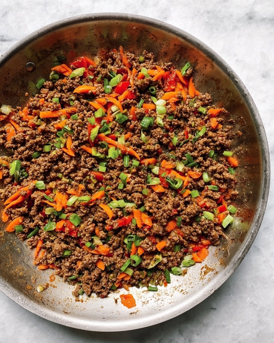 In the image, there is a large stainless steel pan filled with cooked ground beef mixed with small pieces of orange carrot, red bell pepper, and chopped green onions. The beef is brown and crumbly, while the vegetables add bright pops of color scattered evenly throughout. The pan sits on a white marbled surface, showing some cooking marks and a slight shine from the oil on the beef mixture. photo taken with an iphone --ar 4:5 --v 7