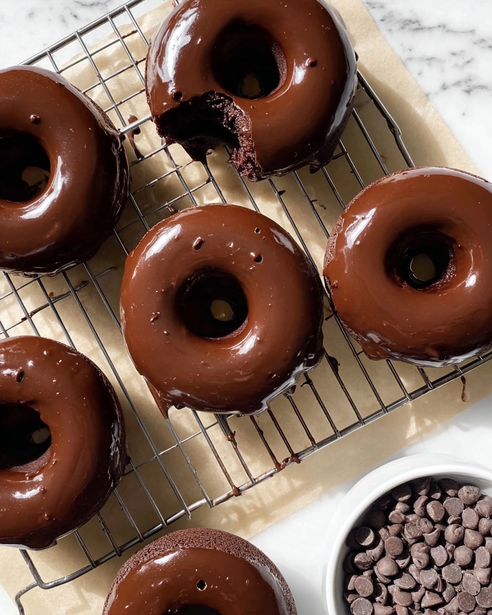 The image shows six chocolate donuts placed on a wire cooling rack over parchment paper, sitting on a white marbled surface. Each donut is covered in smooth, shiny dark chocolate glaze that reflects light softly. One donut near the center has a small bite taken out of it, revealing the soft, dense, dark brown cake inside. The surface of the donuts is evenly glossy, and tiny chocolate drips are visible under the rack. In the lower right corner, part of a white bowl filled with small, round chocolate chips is visible. The lighting is natural, highlighting the rich texture of the chocolate glaze. Photo taken with an iphone --ar 4:5 --v 7