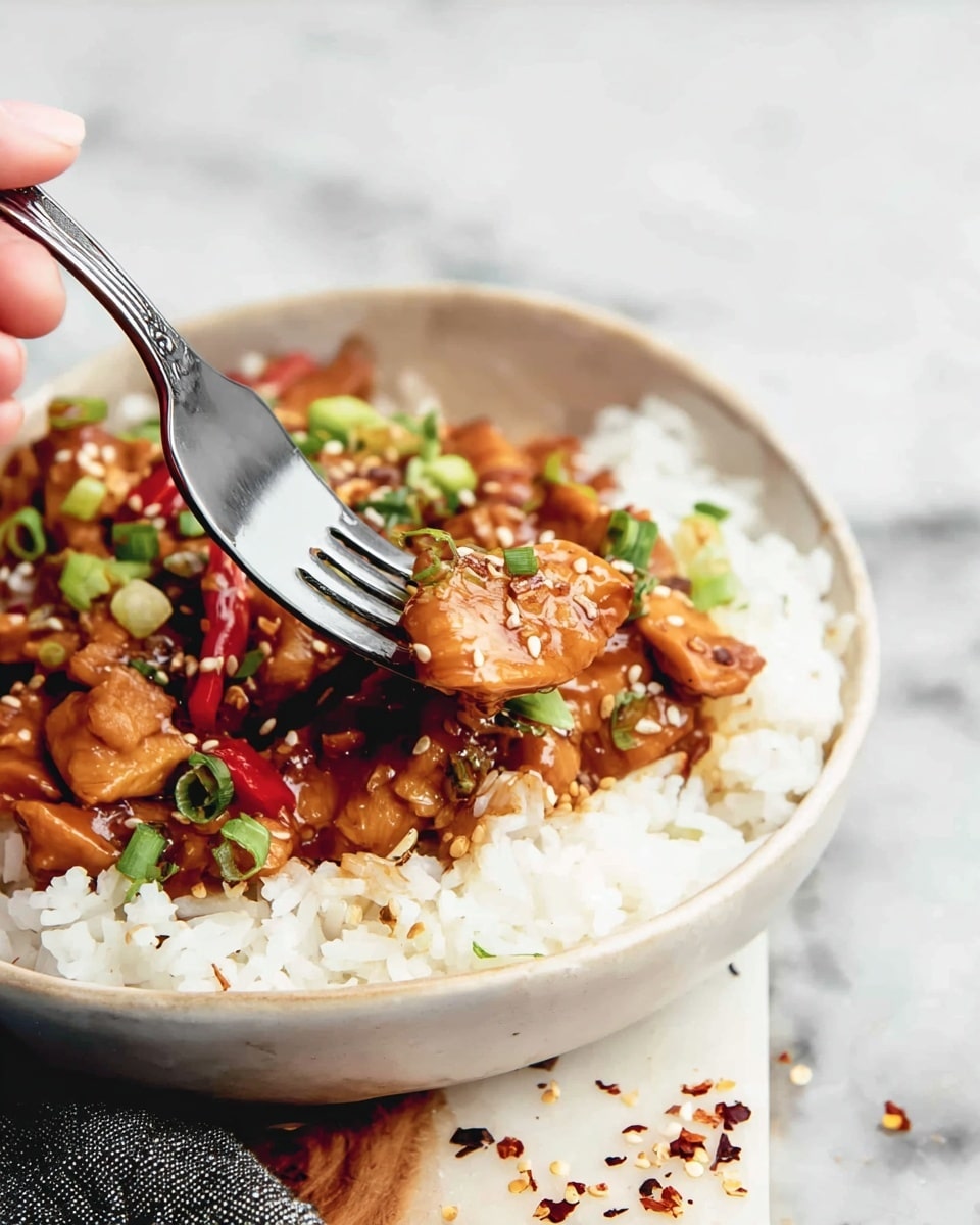 A close-up of a white bowl filled with two layers: the bottom layer is white rice with a soft, fluffy texture, and the top layer features brown pieces of saucy chicken mixed with bits of green onion and red pepper slices. A silver fork is shown lifting a piece of chicken from the bowl, held by a woman's hand. The bowl sits on a white marbled surface with scattered sesame seeds and crushed red pepper flakes around it. photo taken with an iphone --ar 4:5 --v 7