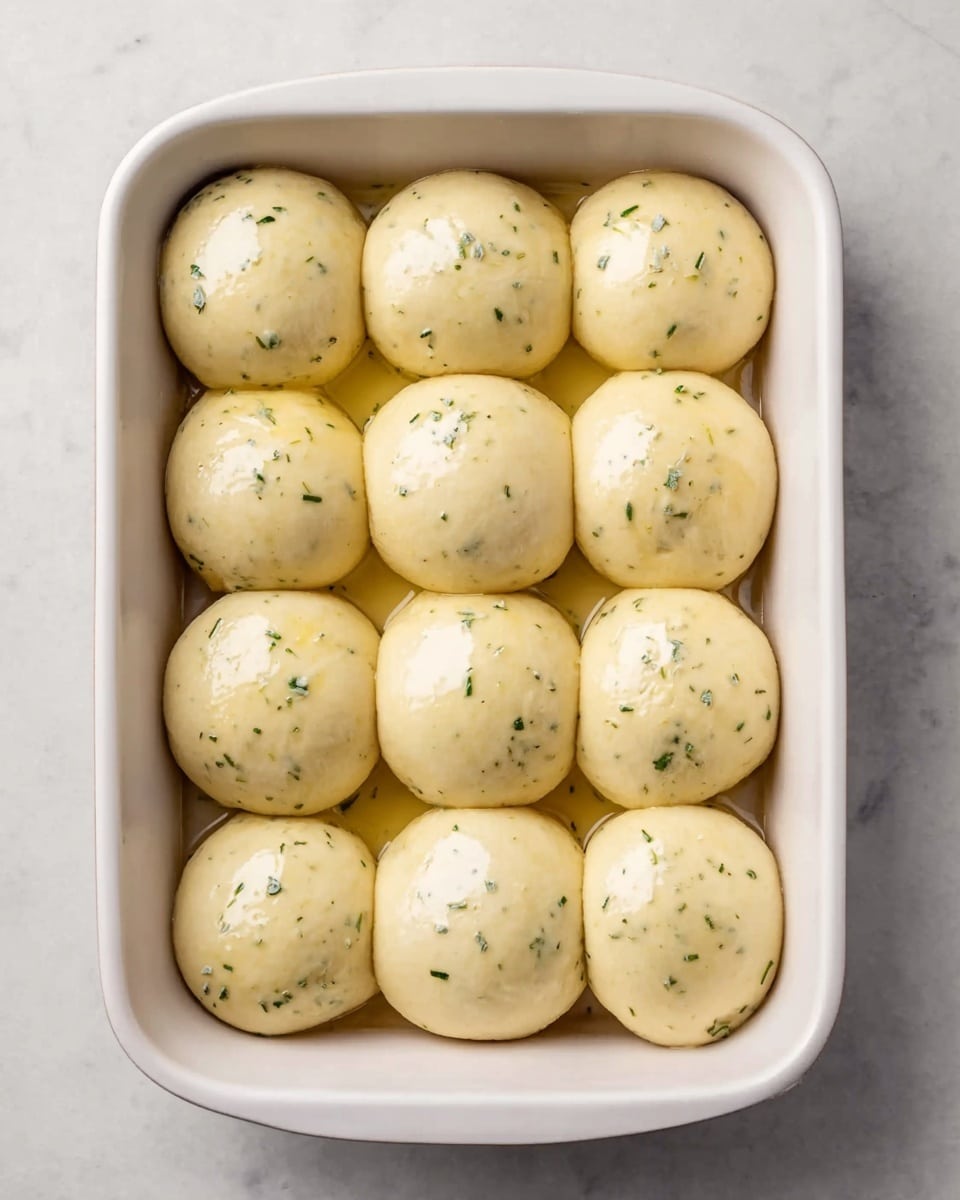 The image shows a white baking dish filled with 12 round, smooth dough balls arranged in a 3 by 4 grid. Each dough ball has small green herb specks evenly spread inside and a shiny surface that suggests they have been brushed with oil or butter. The dough balls are touching each other slightly, filling the dish neatly. The dish is placed on a white marbled surface with soft natural light shining down on the dough, highlighting their smoothness and slight gloss. photo taken with an iphone --ar 4:5 --v 7