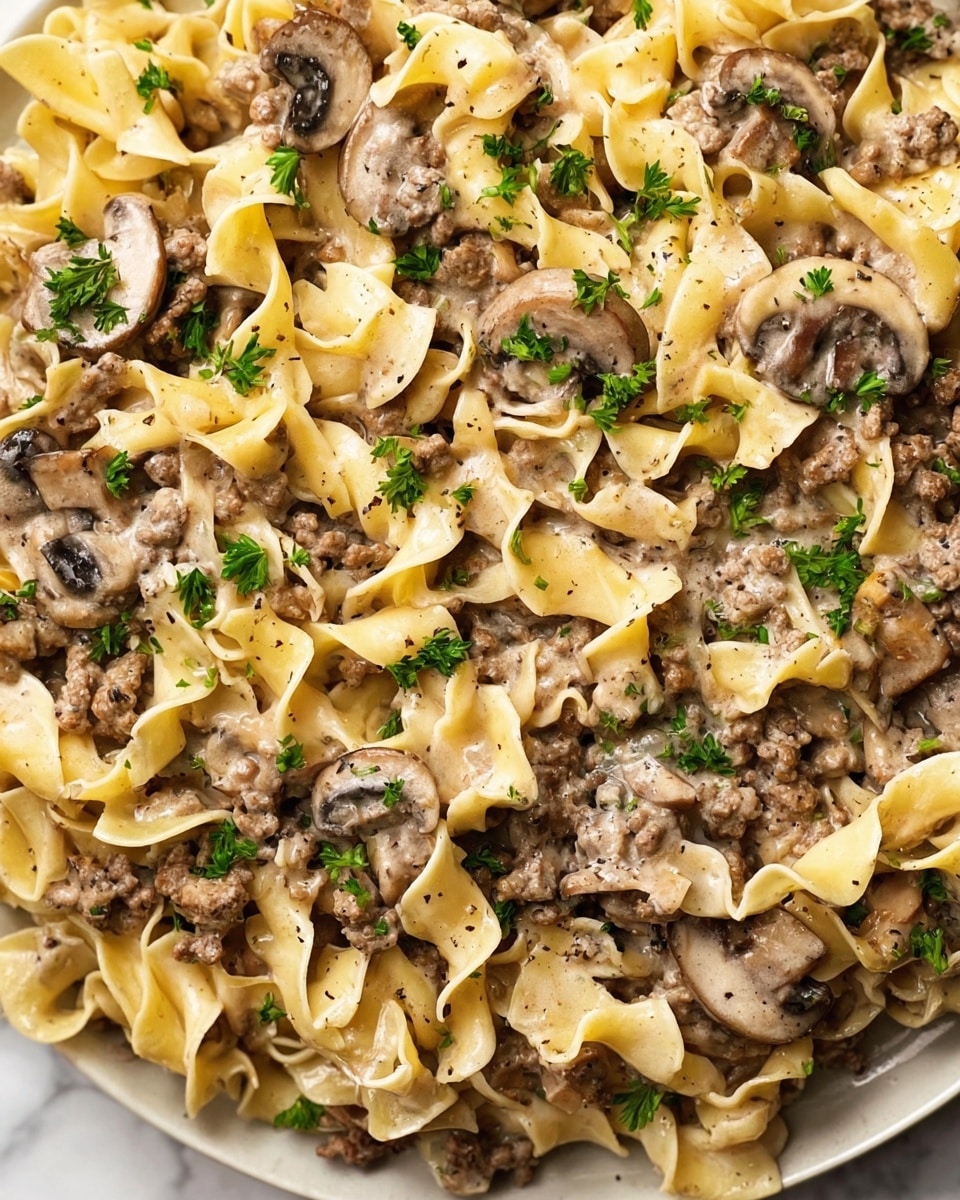 The image shows a close-up of a creamy pasta dish with three main layers: wide, curly egg noodles in a pale yellow color forming the base, browned ground meat scattered throughout the middle layer, and sliced mushrooms that have a slightly golden caramelized look mixed evenly within. Small green parsley leaves are sprinkled on top, adding a fresh color contrast. The creamy sauce lightly coats all the ingredients giving a smooth texture with specks of black pepper adding detail across the dish. The food sits on a white plate placed on a white marbled surface. photo taken with an iphone --ar 4:5 --v 7