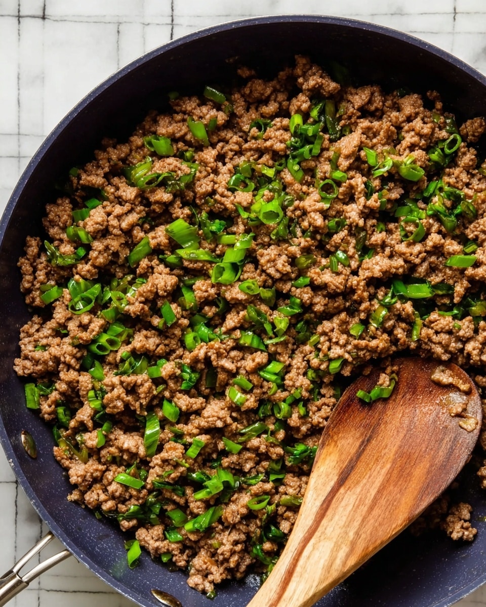 The image shows a close-up view of cooked ground meat mixed with small, bright green chopped scallions scattered evenly throughout. The meat is brown with a cooked texture, filling a dark pan with visible ridges and a wooden spoon resting on the right side, scooping some of the meat. The pan is placed on a white marbled surface with a faint grid pattern underneath. photo taken with an iphone --ar 4:5 --v 7
