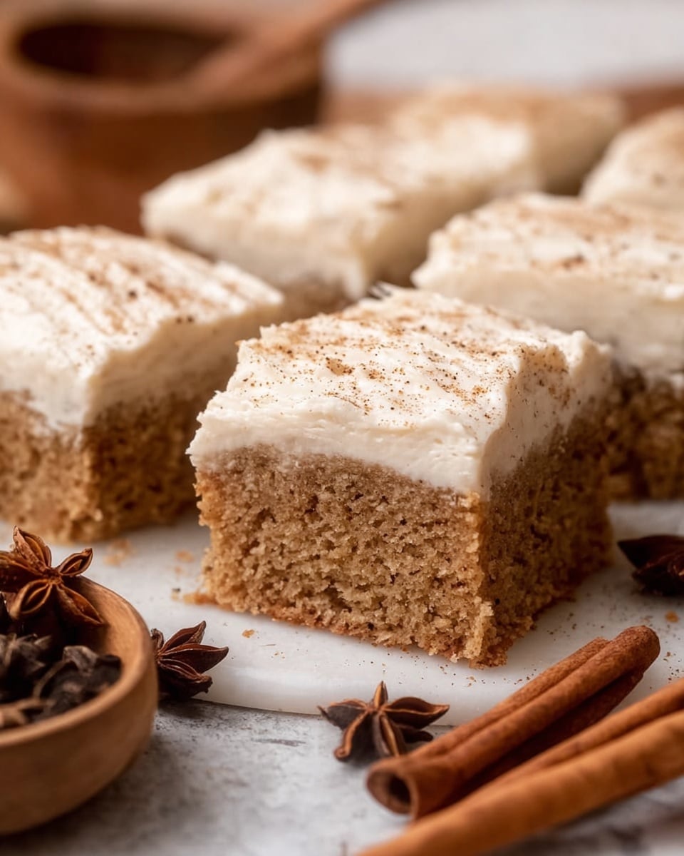 A close-up view of four square pieces of moist spiced cake on a white marbled surface, each piece topped with a thick layer of creamy white frosting sprinkled with a light dusting of ground spice. The cake layer is a light brown color with a soft, crumbly texture. The cakes are arranged closely together in a grid shape. To the left and bottom left of the cakes, there are cinnamon sticks and a wooden bowl containing star anise and other spices. The background is softly blurred, emphasizing the texture and detail of the cake pieces. photo taken with an iphone --ar 4:5 --v 7