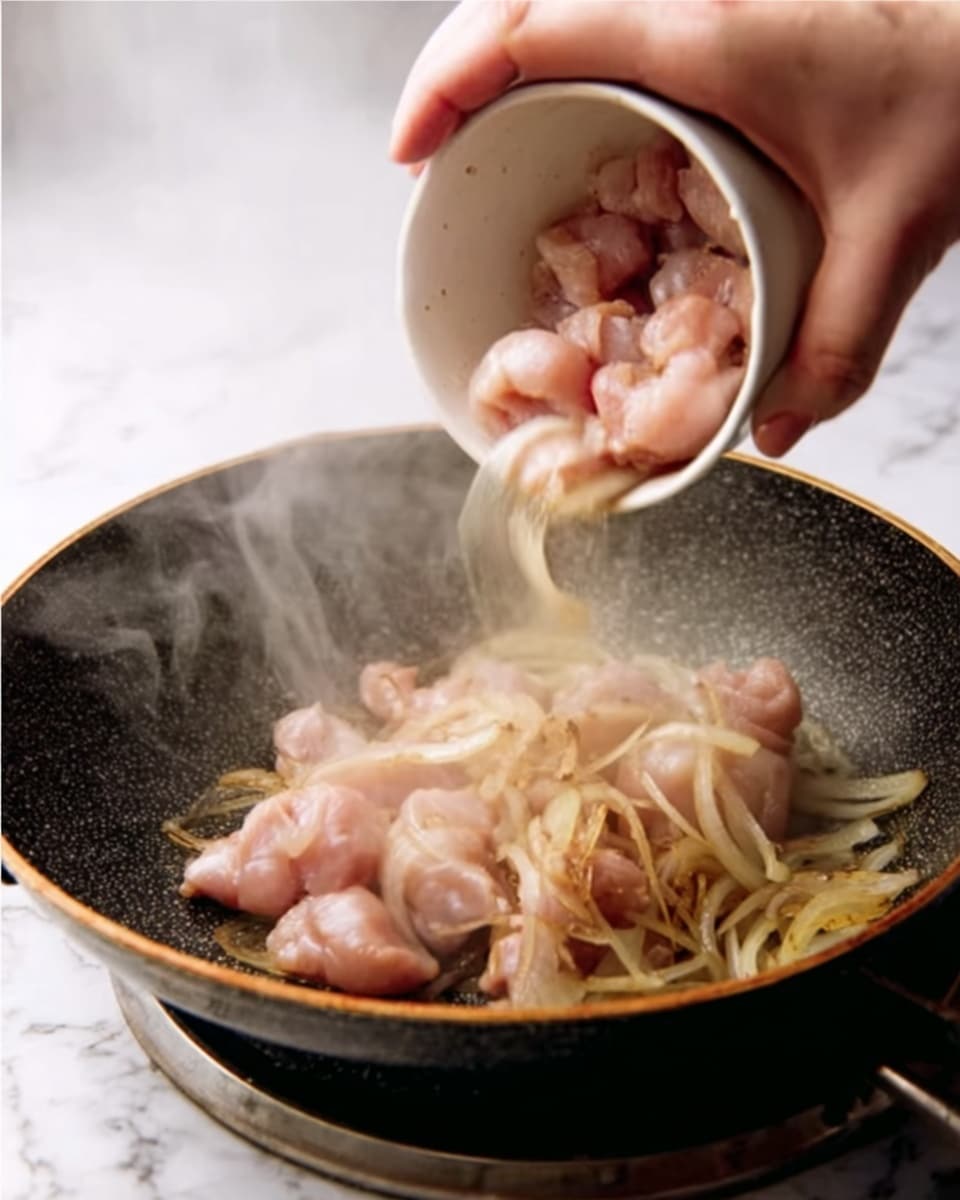 A close-up image shows a woman's hand pouring raw pink chicken pieces from a white bowl into a large dark pan. Inside the pan, there are light brown, translucent sliced onions cooking and some steam rising from the hot pan. The pan has a textured, dark surface and is placed on a white marbled textured background. photo taken with an iphone --ar 4:5 --v 7