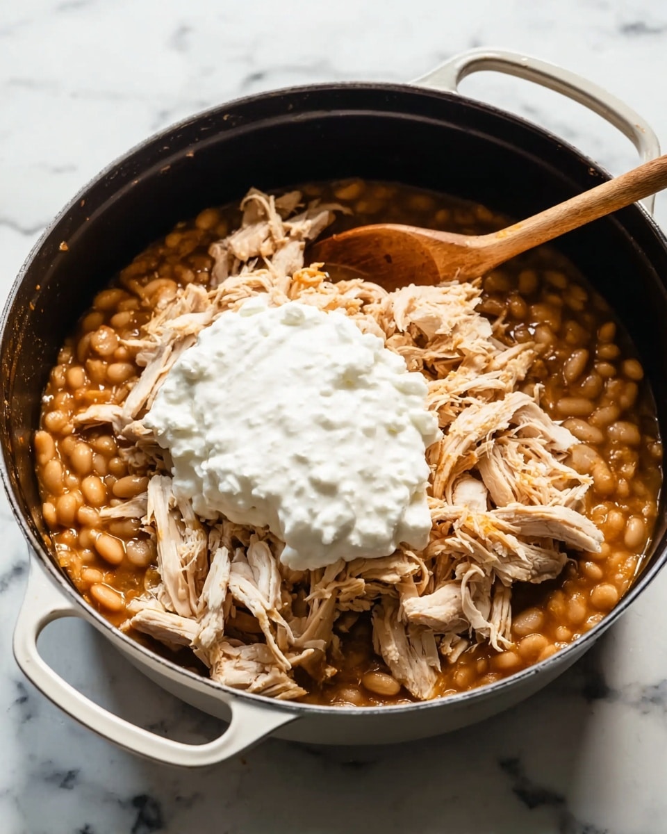 A large black pot with white handles sits on a white marbled surface, filled with a layered dish. The bottom layer is a thick, light brown stew with soft white beans, covering the entire base. On top of this is a generous amount of shredded light brown chicken placed mostly in the center, scattered unevenly. Sitting on the shredded chicken is a heap of thick, creamy white sauce or sour cream, piled high in the middle. A wooden spoon rests inside the pot, its handle pointing out to the top left. Photo taken with an iphone --ar 4:5 --v 7