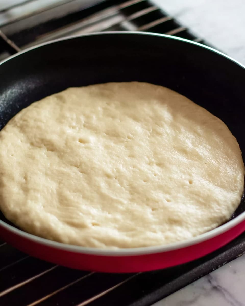A round, pale dough with a smooth and slightly puffy texture sits flat in the center of a black pan. The dough surface looks soft with gentle bubbles forming, showing early signs of cooking. The pan has a shiny black inside and a thin silver rim, while the red side is visible around the edge. The setting is on a stove with metal grates visible in the background, all placed on a white marbled surface photo taken with an iphone --ar 4:5 --v 7