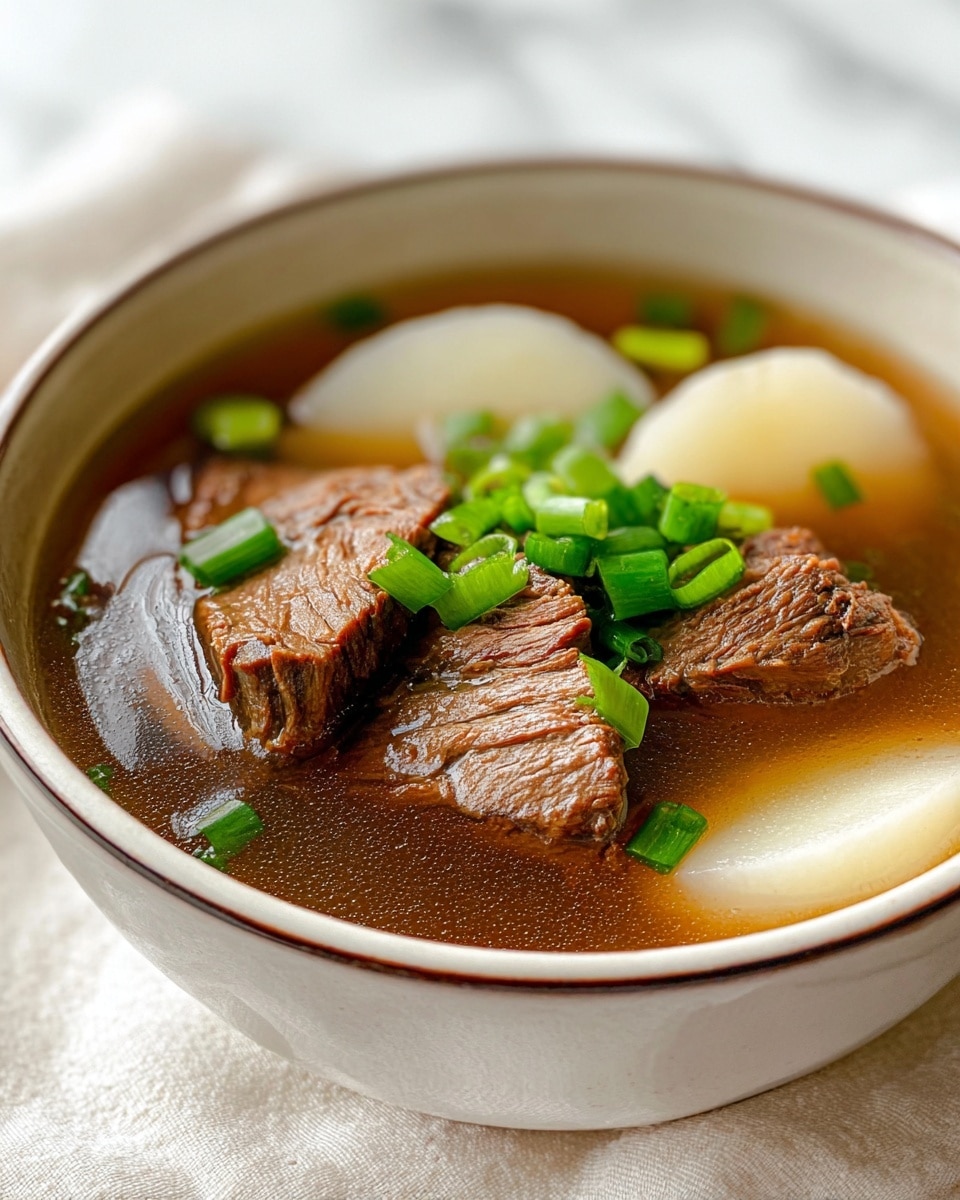 The image shows a bowl of clear broth soup with three pieces of brown, tender-looking meat floating in the light brown liquid. There are two thick, white slices of daikon radish also in the broth. Bright green chopped scallions are sprinkled on top of the soup, adding a pop of color. The bowl is white with a light speckled pattern and is placed on a white marbled surface with a white cloth partly visible next to it. photo taken with an iphone --ar 4:5 --v 7