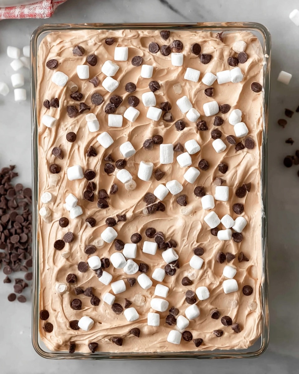 The image shows a rectangular glass dish filled with a thick, creamy layer of light brown mousse or whipped dessert, spread evenly with soft swirls on top. Scattered generously across the surface are small white marshmallows and tiny dark brown chocolate chips, creating a textured contrast on the light brown cream. The dish sits on a white marbled surface, with some of the chocolate chips spilling slightly around the edges. Photo taken with an iphone --ar 4:5 --v 7