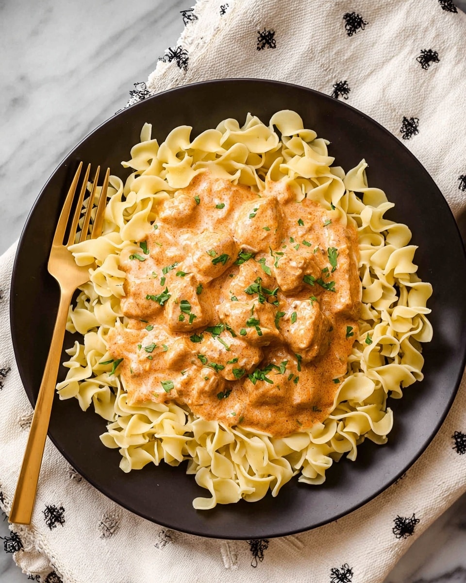 A black plate holds a bed of light yellow egg noodles arranged in a circular shape around the edge, with a creamy orange sauce containing small chunks of meat piled in the center. The sauce is sprinkled with small green parsley leaves for garnish. A gold fork rests on the left side of the plate. The plate is placed on a white marbled surface with a white cloth featuring small black embroidered designs partially visible in the background. photo taken with an iphone --ar 4:5 --v 7