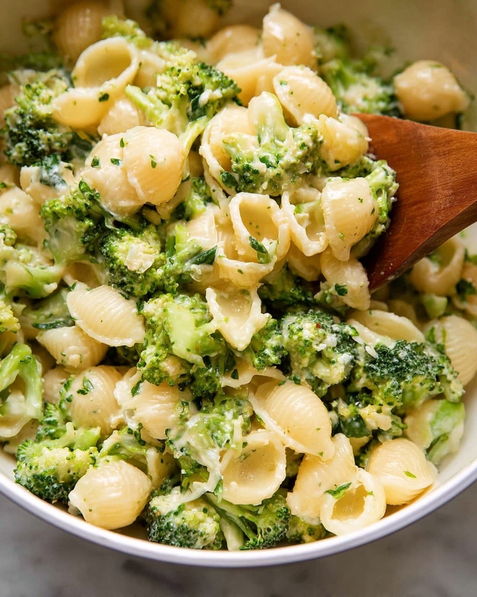The image shows a close-up of a bowl filled with small shell pasta mixed with bright green broccoli florets. The dish has a creamy sauce that lightly coats each piece, giving them a smooth and slightly shiny texture. The shells are pale yellow and mixed evenly with the chopped broccoli, which adds a fresh, green color contrast. A wooden spoon is partially visible on the right side, lifting some of the pasta and broccoli mix. The bowl is white and sits on a white marbled surface. photo taken with an iphone --ar 4:5 --v 7