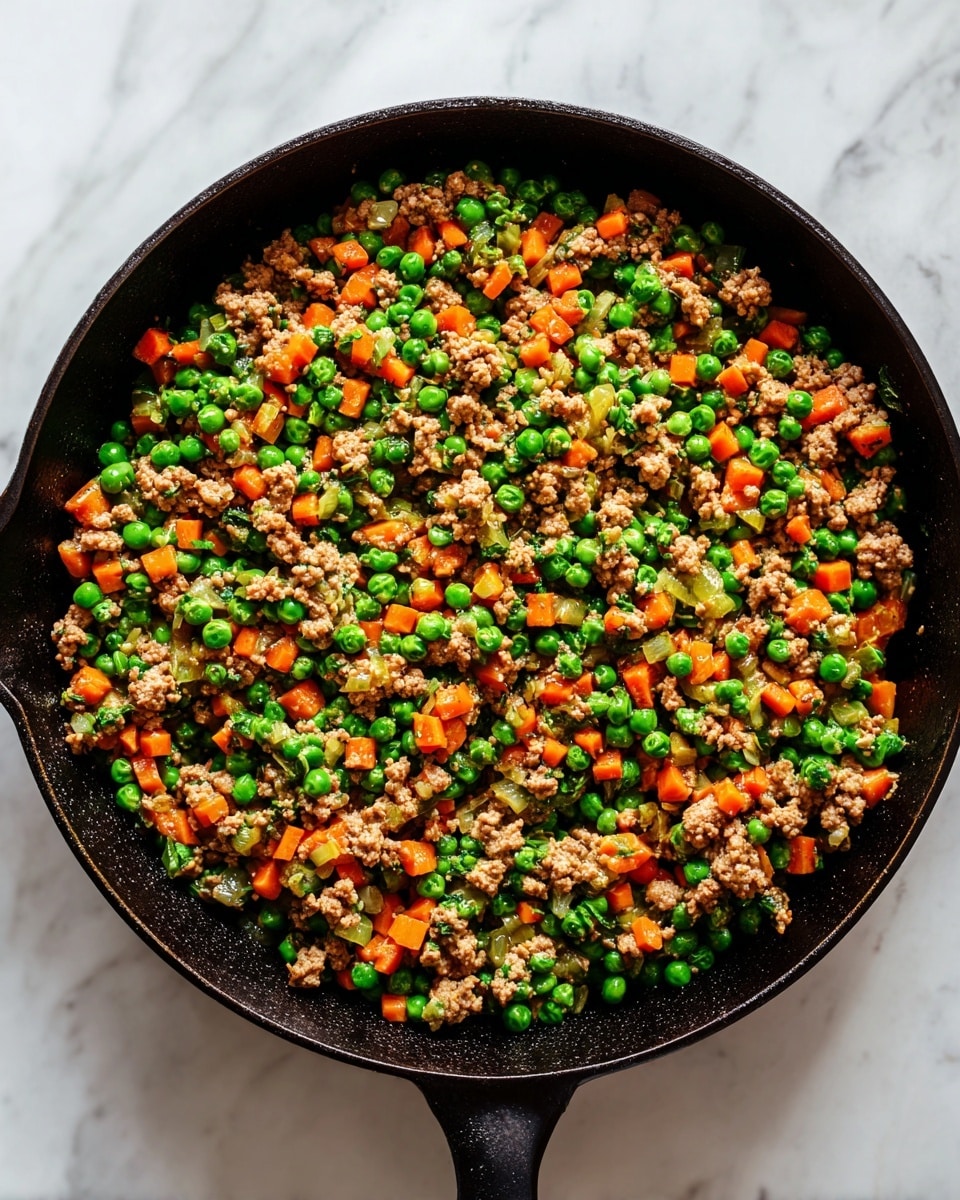 A black cast iron pan filled with a cooked mixture of minced meat, bright green peas, and small orange carrot pieces, all evenly mixed together with a slightly moist texture. The pan rests on a white marbled surface, and the food covers the entire inside of the pan, showing no separate layers, just a uniform combination of ingredients with some bits of onion and seasoning visible. The image focuses on the colorful contrast of the orange, green, and brown colors in the pan. Photo taken with an iphone --ar 4:5 --v 7