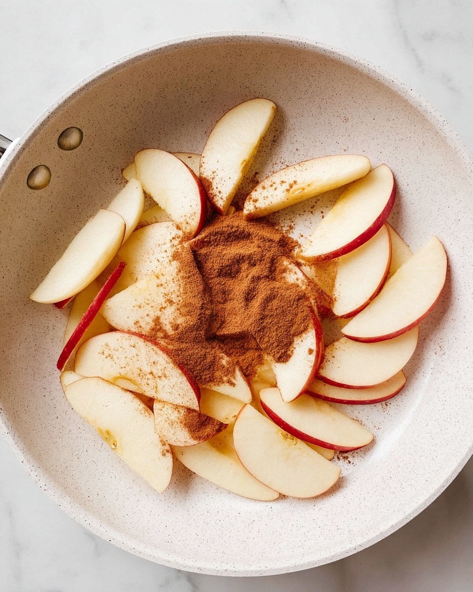 In a white speckled pan, there are many thin apple slices with their red edges showing, arranged in a loose pile across the pan's surface. On top of the apple slices, a small heap of brown cinnamon powder is sprinkled mainly in the center right area, with some powder spreading slightly onto the surrounding slices. The apple slices have a smooth, light cream color with a hint of red along the edges, and the cinnamon powder adds a warm, dusty orange-brown tone. The pan sits on a white marbled surface. photo taken with an iphone --ar 4:5 --v 7