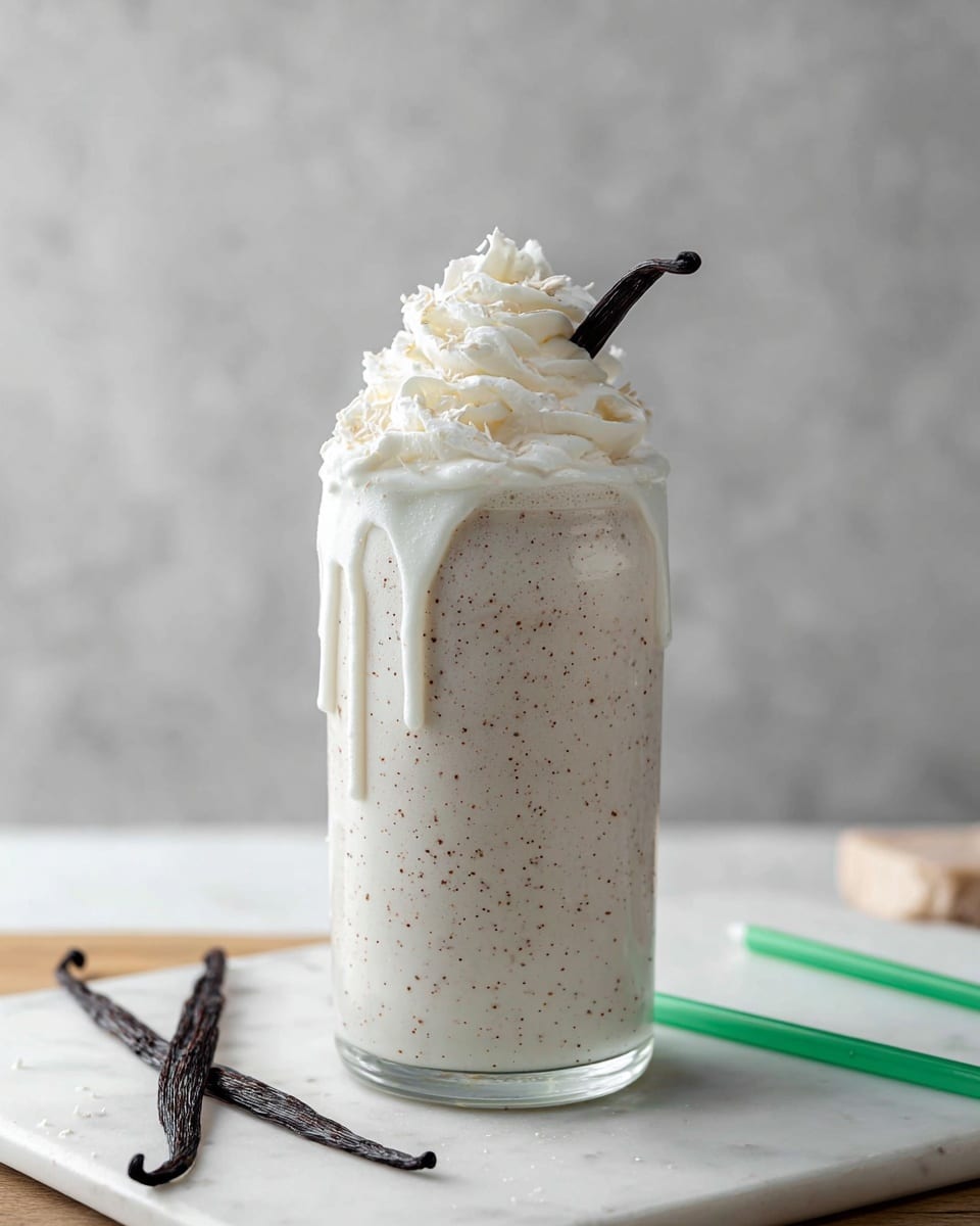 A tall clear glass filled with a creamy white vanilla milkshake that has small brown specks inside. The milkshake is overflowing slightly, with drips running down the sides of the glass. On top, there is a thick swirl of white whipped cream garnished with a small dark vanilla bean pod. The glass sits on a white marbled surface with two dark vanilla bean pods lying nearby, and a green straw placed in front. The background is a soft gray with a subtle texture. Photo taken with an iphone --ar 4:5 --v 7