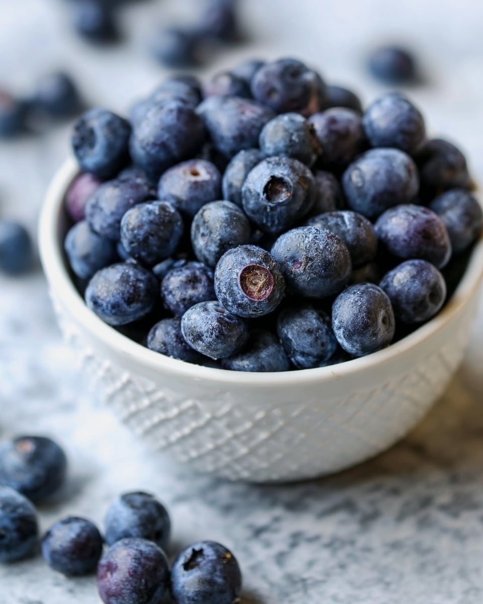 A close-up image of a white bowl filled to the top with fresh blueberries, each berry showing a deep blue to purple color with a natural matte texture. A few blueberries are scattered loosely around the bowl on a surface with a white marbled texture. The focus is sharp on the berries inside the bowl, showing details of their round shape and slight natural imperfections. The bowl itself has a small raised diamond pattern on its outside and is centered in the frame. photo taken with an iphone --ar 4:5 --v 7