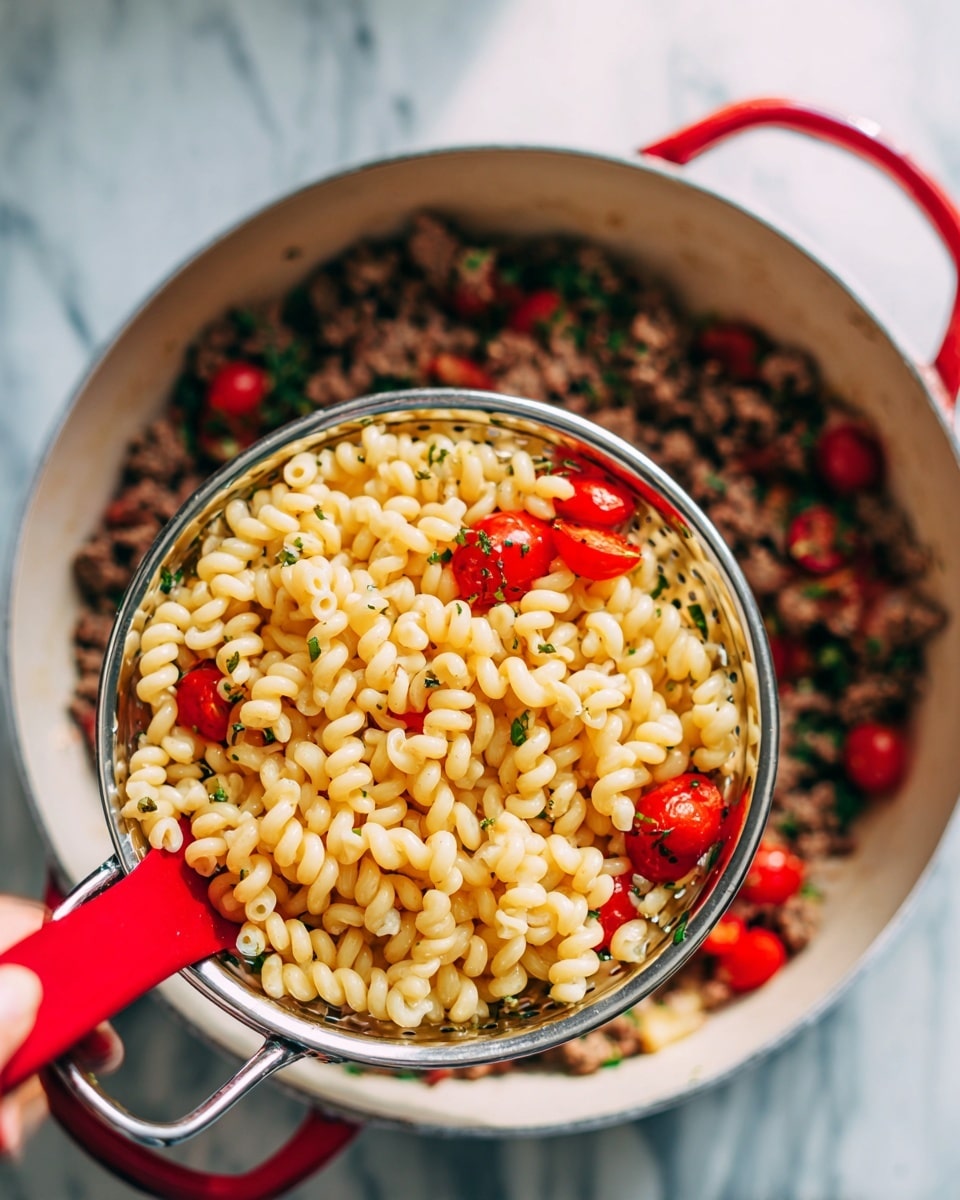 A close-up image shows a woman’s hand holding a silver colander filled with cooked curly elbow pasta, which is a light yellow color. The colander is positioned over a large white pot with a red outside, containing a mix of browned ground meat, halved red cherry tomatoes, and green herbs evenly spread out. A red spoon rests inside the pot amid the ingredients. The background panel is a white marbled surface, and soft natural light brightens the scene. Photo taken with an iphone --ar 4:5 --v 7