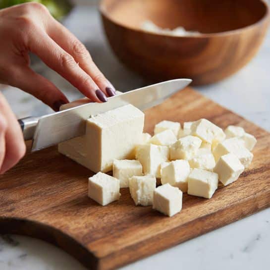 A close-up image shows a woman's hand holding a knife and slicing a block of white cheese into small cubes on a wooden cutting board. The cheese has a smooth texture and clean edges. Several cheese cubes are already cut and placed next to the block on the board. The background includes a wooden bowl and a blurred setting behind a white marbled surface. Photo taken with an iphone --ar 4:5 --v 7