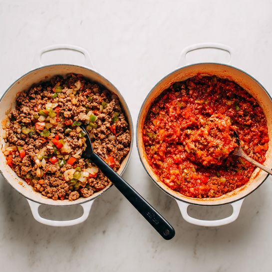 The image shows two white pots, each filled with different stages of cooking ground meat mixed with vegetables. The pot on the left contains cooked ground meat combined with chopped white onions and small diced pieces of green and red bell peppers, all lightly browned and textured with small and medium bits; a black spoon is stirring the mixture on the right side of the pot. The pot on the right holds a saucier mixture where the browned ground meat is now combined with a chunky, vibrant red tomato sauce containing visible tomato pieces and softened bell peppers and onions, with the black spoon lifting some of the thick, textured sauce in the center. The pots sit on a white marbled surface. photo taken with an iphone --ar 4:5 --v 7