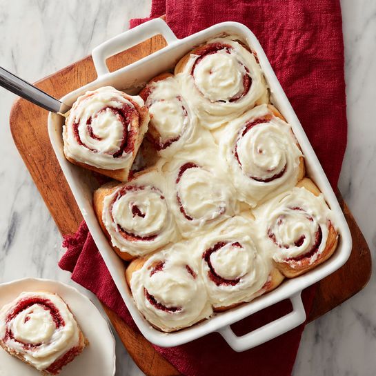 A white rectangular baking dish holds a batch of cinnamon rolls covered with thick, creamy white icing. The cinnamon rolls are arranged in three rows and three columns, with parts of three rolls missing from the top left corner where some rolls have been taken out, showing a brown spiral pattern underneath the icing. The rolls have a reddish-brown color with swirls visible beneath the smooth, slightly melted white icing layer. The baking dish sits on a white marbled surface. Around the dish are two silver forks on the right, a gray-white plate with a cinnamon roll slice on the bottom left, and some small heart-shaped red paper cutouts scattered on the surface. In the background, a sheer red cloth is partially visible on the left side. A clear glass bowl with strawberries is on the upper right corner. Photo taken with an iphone --ar 4:5 --v 7