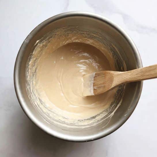 A close-up view inside a silver mixing bowl showing a creamy, light brown batter with a smooth and thick texture. A wooden spatula with batter coating its edge rests on the right side inside the bowl. The bowl sits on a white marbled surface. The lighting is soft and natural, highlighting the batter's glossy and soft appearance. photo taken with an iphone --ar 4:5 --v 7