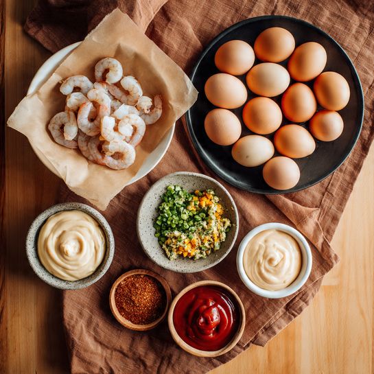 The image shows several bowls and a plate with ingredients arranged on a wooden surface with a brown cloth underneath. In the top left, there is a white plate lined with brown parchment paper holding raw shrimp, which are pale pink and curled. Below it to the left, a small gray bowl has chopped green and yellow vegetables that look fresh and finely diced. Towards the bottom left, a small white bowl filled with smooth, creamy mayonnaise is visible. On the right side, a larger black plate holds a dozen light brown eggs piled neatly. Above the eggs, a small stone bowl contains reddish-brown seasoning with visible herbs, and next to it, a small white bowl with two kinds of sauce, one bright red and the other creamy beige with a grainy texture. The whole layout sits on a white marbled surface. photo taken with an iphone --ar 4:5 --v 7