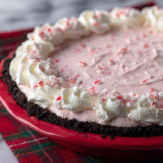 The image shows a peppermint cream pie with three visible layers. The bottom layer is a dark, crumbly chocolate crust tightly packed in the edge of a red pie dish. The middle layer is a smooth, pale pink cream with gentle swirls in the center, speckled with small crushed pieces of red and white peppermint candy. The top layer is a thick border of fluffy white whipped cream piped around the edge, also sprinkled with crushed peppermint candy. The pie sits on a red and green plaid cloth on a white marbled surface. photo taken with an iphone --ar 4:5 --v 7