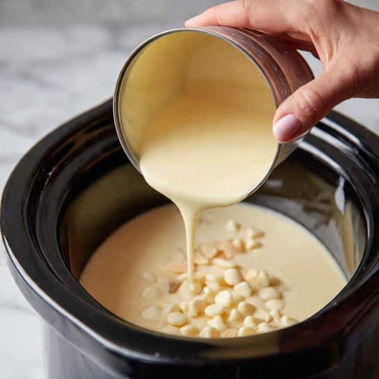 A close-up image shows a woman's hand pouring smooth, thick, pale yellow sweetened condensed milk from a can into a slow cooker filled with coconut milk and small white chocolate chips or butter bits visible on the surface. The slow cooker has a dark rim, and the liquid inside looks creamy and glossy. The background surface is a white marbled texture. Photo taken with an iphone --ar 4:5 --v 7