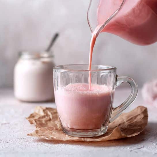 A clear glass mug sits on crumpled brown paper on a white marbled surface, while a soft pink liquid is being poured into it from a matching pink jug. The pink liquid has a smooth texture and fills the glass nearly halfway. In the background, there is a blurred glass jar with a white creamy substance and a spoon inside. The overall scene has a calm and fresh feel, with the clear glass showing the layers of the pink drink. Photo taken with an iphone --ar 4:5 --v 7