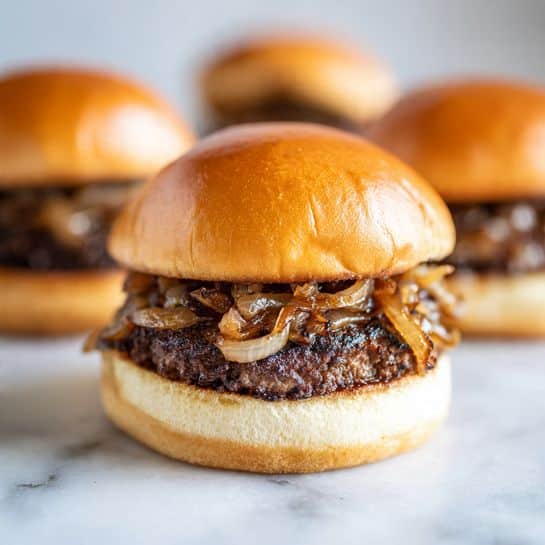 The image shows a close-up of a small burger with three visible layers, placed on a white marbled surface. The bottom layer is a soft, light beige bun with a smooth texture. On top of that lies a thick, juicy, dark brown beef patty with a slightly crumbly surface. Above the beef patty is a generous layer of caramelized onions, glossy and golden brown with some translucent parts. The top layer is a shiny, golden-brown bun, slightly domed and smooth, completing the burger. The background is softly blurred, showing hints of more burgers. photo taken with an iphone --ar 4:5 --v 7
