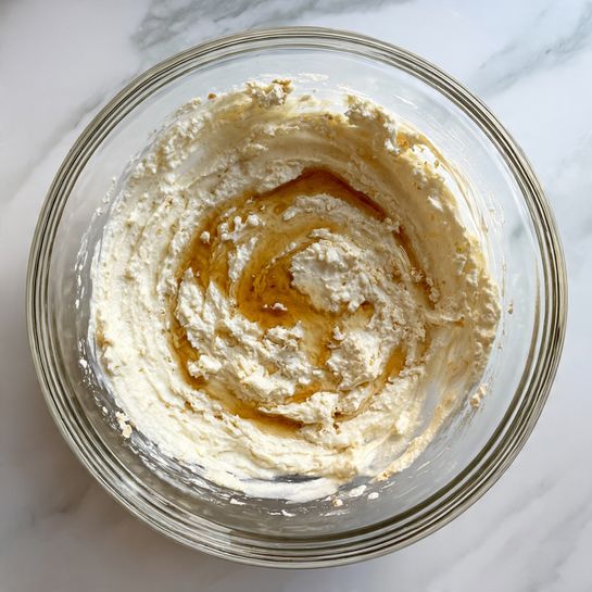 A clear glass bowl holds a thick, creamy mixture of off-white batter with visible clumps, surrounding a small pool of golden-brown liquid in the center. The batter has a slightly grainy texture, spread unevenly along the inner sides of the bowl, which sits on a white marbled surface. photo taken with an iphone --ar 4:5 --v 7