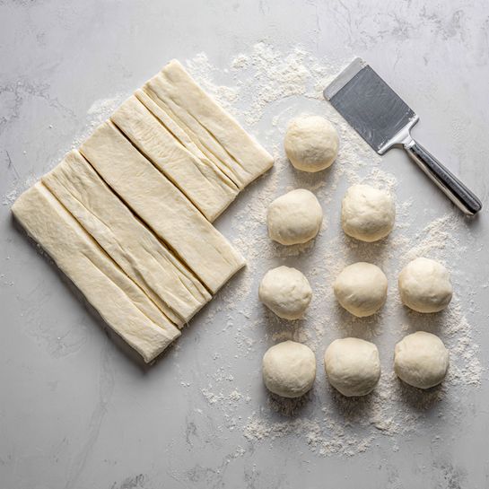 Cheese Danish Recipe 4 The image shows two side-by-side pictures of dough pieces on a light grey surface with a metal dough scraper placed near the edges. On the left side, there are about twelve rectangular dough pieces with smooth, layered textures and soft edges. On the right side, there are twelve smooth round dough balls, evenly shaped, with a slightly floury surface and soft creases. The surface has a subtle white marbled texture throughout. photo taken with an iphone --ar 4:5 --v 7
