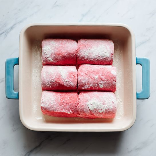 Four pieces of red dough layers lie flat on a white marbled surface. The dough has a dusting of white flour giving it a soft texture and a slightly wrinkled appearance. Behind the dough pieces, there is an empty rectangular white baking dish with light pink inside and blue sides. The dish has two handles on each end. Photo taken with an iphone --ar 4:5 --v 7