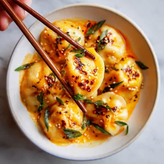 A close-up of a dumpling held by wooden chopsticks in a woman's hand, showing its smooth, light orange dough with a slightly shiny texture from a creamy, reddish-orange sauce. The dumpling is topped with small dark chili oil pieces and sesame seeds, with tiny green herb bits scattered on it. Below, many similar dumplings are partially visible in a white dish, covered in the same creamy sauce with green herbs sprinkled around. The background has a soft focus white marbled texture. photo taken with an iphone --ar 4:5 --v 7