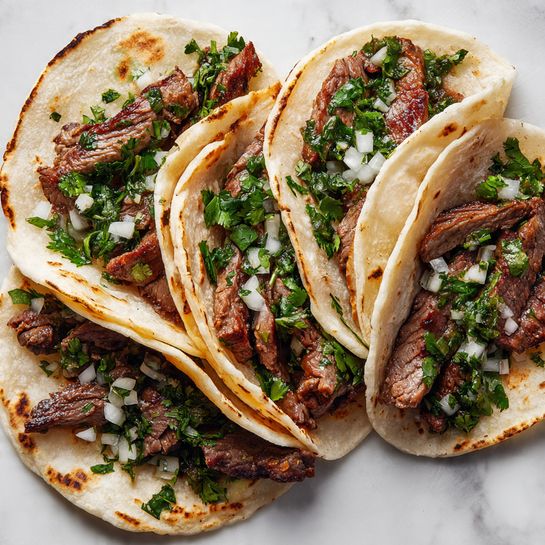 Four small soft corn tortillas, each filled with dark brown grilled beef pieces, topped with light green chopped fresh cilantro and small cubes of white and purple onion. The tortillas show some browned grilled spots and are placed close together on a white marbled surface. In the background, the top half of a green lime and some cilantro leaves are visible, adding a fresh touch. The overall look is bright and colorful with a rustic, fresh feel photo taken with an iphone --ar 4:5 --v 7