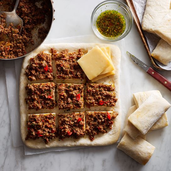 The image shows a white rectangular dough sheet divided into six squares on a white marbled surface. Two of the top squares each have a layer of browned ground meat with visible seasoning and small red bits, while the left square also has two rectangular slices of marbled yellow and white cheese on top of the meat. To the left, a pan filled with extra cooked ground meat and a spoon is partly visible. On the right side, a baking sheet with parchment paper and a glass bowl containing a green sauce sit above the dough. Nearby, a knife rests next to additional slices of the same marbled cheese. Photo taken with an iphone --ar 4:5 --v 7