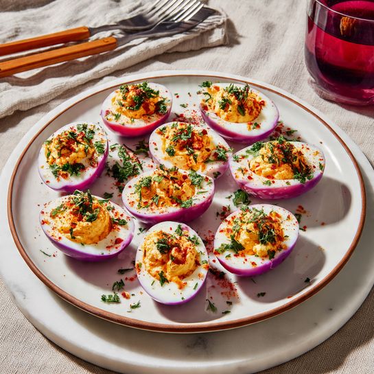 There are eleven halved eggs arranged on a round white plate with a brown rim, placed on top of a white marbled round tray. Each egg has a vibrant purple outer white layer, a white inner white layer, and a creamy pale orange filling with small green herb pieces. The filling is slightly sprinkled with red spice and fresh chopped green herbs. The plate sits on a light beige cloth with two forks that have wooden handles on the right side and a dark red drink in a clear container in the background. The photo taken with an iphone --ar 4:5 --v 7