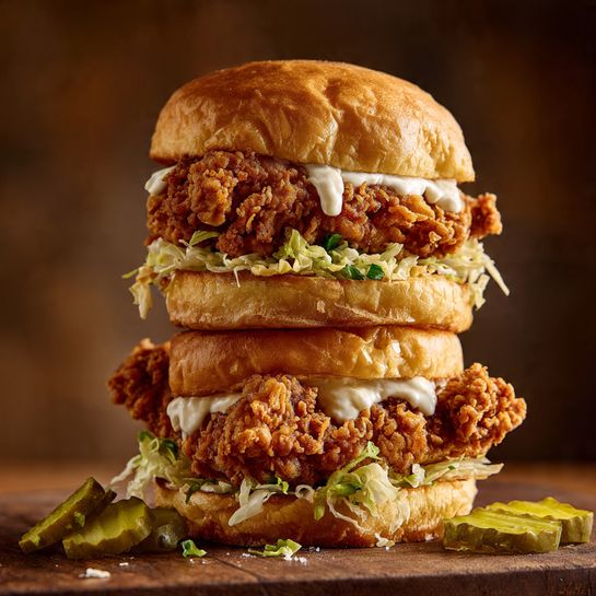 The image shows a double fried chicken sandwich stacked tall with two soft, golden brown buns on top and at the bottom. Inside, each bun holds a crispy, richly golden fried chicken fillet with a crunchy texture. Between the layers, there is a fresh, shredded layer of white lettuce that peeks out and some creamy white sauce spread on the inside of the buns. Thin green pickle slices rest under the chicken pieces, adding a slight color contrast. The sandwich is set on a wooden surface with a few stray pickles and lettuce shreds around it, with a blurred brown background enhancing the warm tones. photo taken with an iphone --ar 4:5 --v 7