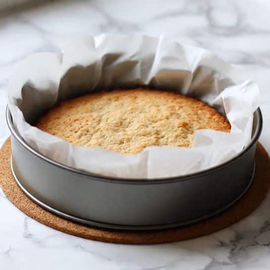 The image shows a round baked cake with a slightly golden brown top, inside a metal tart tin with fluted sides. The cake is lined with white parchment paper that extends above the sides of the tin, showing soft folds and crinkles. The tin sits on a round cork mat on a white marbled surface. The cake looks soft and lightly textured on top, with no decorations or layers visible. Photo taken with an iphone --ar 4:5 --v 7