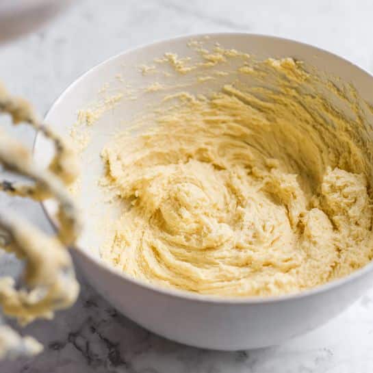 A white bowl filled with light yellow, soft and creamy dough with a slightly rough texture, showing small swirls and peaks on top. The dough spreads evenly inside the bowl, with some stuck on the sides. In the foreground, a blurred metal mixer beater with bits of dough clinging to it is visible. The bowl sits on a white marbled surface. photo taken with an iphone --ar 4:5 --v 7