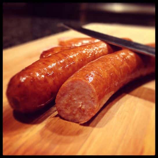 Two shiny brown sausages lie on a wooden cutting board with one sausage being sliced thinly by a knife held below the frame. The sausages have a smooth, slightly glossy texture with some small wrinkles. A thin, black chopstick is placed under the sausage being sliced, showing careful cutting. The background is out of focus but appears warm-toned. photo taken with an iphone --ar 4:5 --v 7