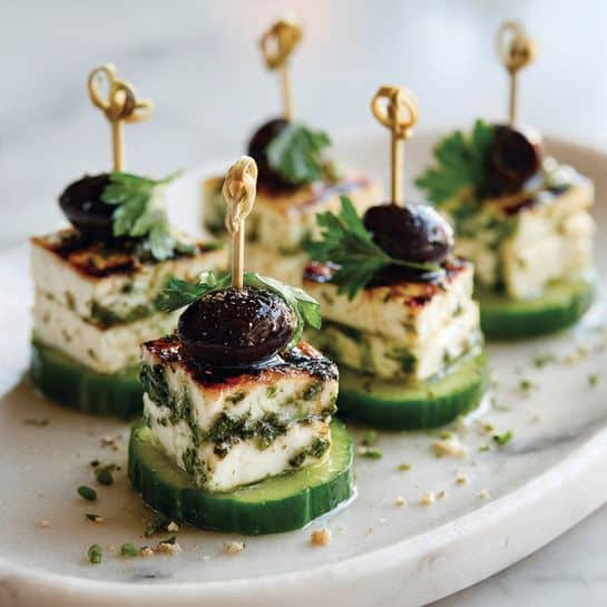 The image shows five small stacked appetizers on a white plate with a white marbled surface underneath. Each appetizer has three layers: a round green cucumber slice at the bottom with a slightly shiny and fresh texture, a thick square of grilled feta cheese in the middle that is white with green herbs and light brown grill marks on top, and a shiny black olive on top. Each piece is held together with a small wooden skewer that also has a small green parsley leaf stuck under the olive, adding a fresh and colorful detail. The appetizers are arranged in a neat row, with some small bits of seasoning or herbs scattered on the plate. photo taken with an iphone --ar 4:5 --v 7