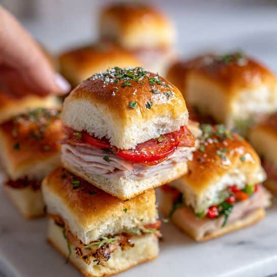 The image shows a close-up of a sandwich slider being lifted by a metal spatula over a white marbled surface, surrounded by several other sliders. The slider has three visible layers starting with a soft, golden brown bread roll on the bottom, followed by thin layers of pink deli meat stacked neatly, a layer of bright red roasted peppers, a few slices of light yellow cheese, and topped with a shiny golden brown bread roll sprinkled with green dried herbs. The bread looks soft with a slightly crispy crust on top. The overall look is warm and fresh. photo taken with an iphone --ar 4:5 --v 7